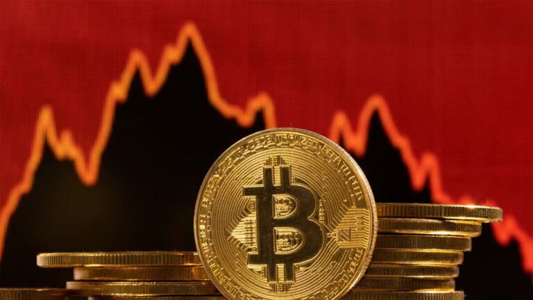 Abwärts zeigender roter Kurs-Chart hinter einer physischen Bitcoin-Münze auf einem Schreibtisch.