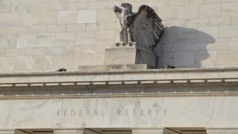 Ein Bitcoin-Symbol vor einem verschwommenen Kurs-Chart und dem Gebäude der US-Notenbank Federal Reserve