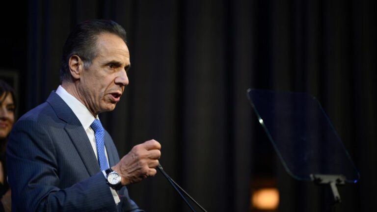 Andrew Cuomo spricht vor Mikrofonen über Regulierung, im Hintergrund verschwommene Kurscharts von Bitcoin und Ethereum